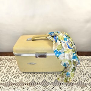 May include: A vintage, pale yellow suitcase with a tan handle and silver trim. A floral scarf in shades of blue, green, and white is draped over the top. The suitcase is sitting on a lace tablecloth.