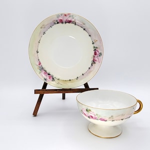 May include: A vintage teacup and saucer set with a delicate floral design. The saucer features a ring of pink roses and green leaves around the edge, while the teacup has a similar design around the rim. Both pieces are white porcelain with a gold trim.