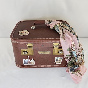 May include: A brown hard-shell suitcase with gold-tone metal latches and a top handle. The exterior features three stickers depicting Paris landmarks and a pink floral patterned scarf draped over the right side of the case. The case has white stitching.