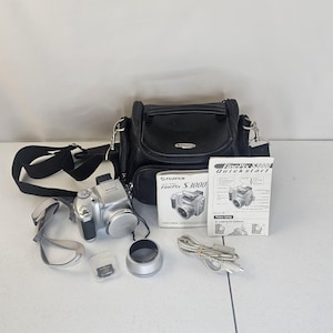 May include: A silver Fujifilm FinePix S3000 digital camera with a black camera bag, lens cap, memory card, and charger cable. The camera has a strap and a quick start guide.