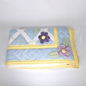 May include: A folded blue quilt with a floral design. The quilt features a light blue background with a diamond pattern, yellow borders, and appliqued purple and pink flowers with yellow centers. Green leaves are also appliqued.