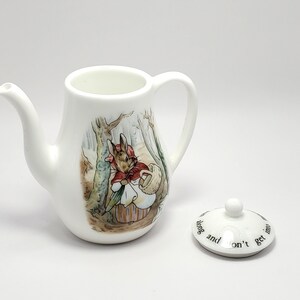 May include: A white ceramic teapot with a Peter Rabbit illustration and a matching lid. The lid has the text "Don't get into mischief" on it.