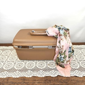May include: A vintage brown suitcase with a handle and silver accents. A pink scarf with a floral pattern is draped over the top. The suitcase is on a wooden surface with a white lace runner.