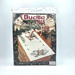 May include: A Bucilla brand embroidery kit in a clear plastic package. The kit features a Christmas table runner design with doves, holly, and the text "Doves and Holly". The table runner measures 18" x 44".