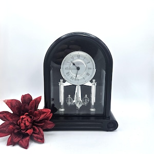 May include: A black mantel clock with a silver face and Roman numerals. The clock has a glass front and a pendulum with three glass orbs. A dark red flower is in the foreground.