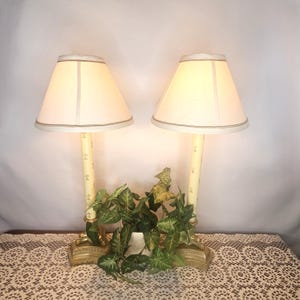 May include: A pair of cream-colored table lamps with off-white shades. The lamp bases are a light yellow with a fleur-de-lis pattern. The lamps are on a gold-colored base, and there is a small plant between them.