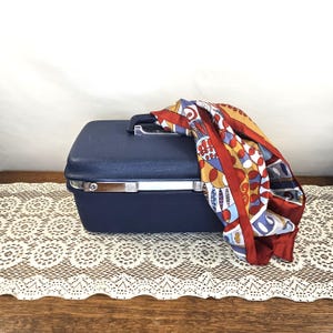 May include: A navy blue vintage suitcase with silver hardware and a black handle. A colorful silk scarf with red, yellow, and blue patterns is draped over the top. The suitcase rests on a cream-colored lace doily.