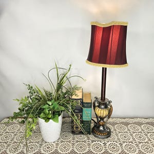 May include: A decorative table lamp with a burgundy shade and gold trim. The lamp has a dark brown stem and a detailed base. A white textured pot with greenery and two vintage books are next to the lamp.