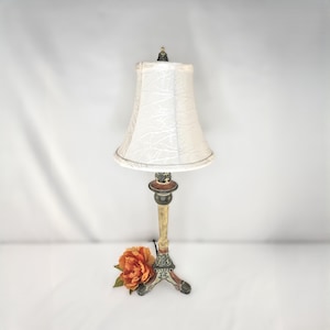 May include: A table lamp with a white, textured, bell-shaped shade and a slender, multi-colored base featuring black, gold, and red tones. The base has a tripod-style footed design and is positioned next to a single orange-colored flower.