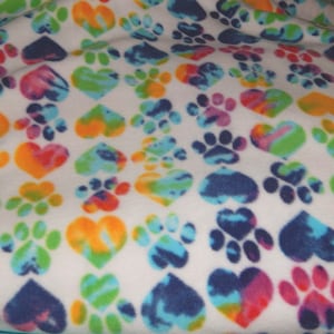 May include: White fleece fabric with a repeating pattern of colorful tie-dye hearts and paw prints. The hearts and paw prints are in shades of orange, yellow, blue, green, and purple.
