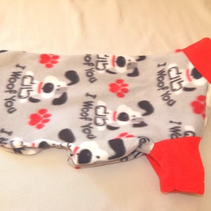 Op de afbeelding: Grijze honden onesie met een herhaald patroon van cartoonhonden, rode pootafdrukken en de tekst "I Woof You". De mouwen en de boorden zijn felrood. Een gezellig outfit voor een kleine hond.