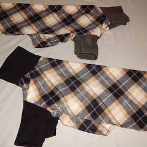 Op de afbeelding: Twee hondenjassen met een crème, zwart en tan ruitpatroon. De mouwen en halsbanden zijn effen donkergrijs of zwart. De jassen zijn gemaakt van een zacht, fleece-achtig materiaal.