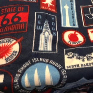 Op de afbeelding: Een blauwe fleece deken met een patroon van kleurrijke staatspatchen. De patches tonen staatsnamen, bezienswaardigheden en symbolen. De patches zijn in een willekeurig patroon gerangschikt.