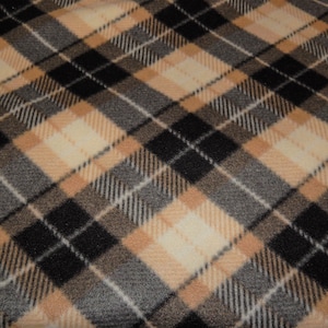Op de afbeelding: Een close-up van een geruite stof met een herhalend patroon van zwarte, bruine, crème en grijze diagonale lijnen. De stof heeft een zachte, getextureerde uitstraling, wat suggereert dat het een deken of een soortgelijk textiel kan zijn.