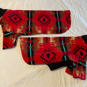 Op de afbeelding: Twee hondenjassen met een patroon geïnspireerd op het zuidwesten. De jassen hebben een rode achtergrond met geometrische ontwerpen in zwart, bruin en turquoise. De mouwen en halslijnen zijn zwart.