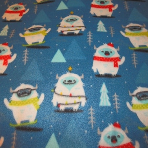 Op de afbeelding: Blau fleece stof met een herhalend patroon van witte yeti-personages. Sommige yeti's dragen rode sjaals, mutsen of zonnebrillen. Anderen zijn versierd met kerstverlichting of skiën op groene ski's. De achtergrond heeft lichtblauwe driehoeken en dennenbomen.