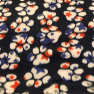 Op de afbeelding: Een marineblauwe fleece deken met een herhalend patroon van witte poot afdrukken. Elke poot afdruk heeft rode en blauwe sterren erin.