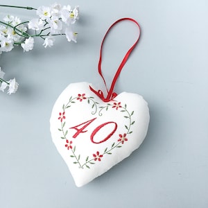 May include: White heart-shaped cushion with red embroidery of a floral wreath and the number "40" in the centre. The cushion has a red ribbon loop for hanging.