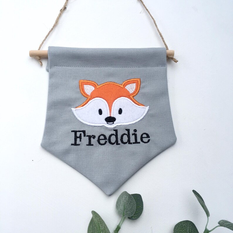 May include: A gray fabric wall hanging with a fox design and the name "Freddie" embroidered in black. The fox is orange and white, and the banner is suspended by a wooden dowel and twine.