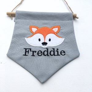 May include: A gray fabric banner with a fox design and the name "Freddie" embroidered in black. The fox has orange and white details. The banner is suspended by a wooden dowel and a natural twine.