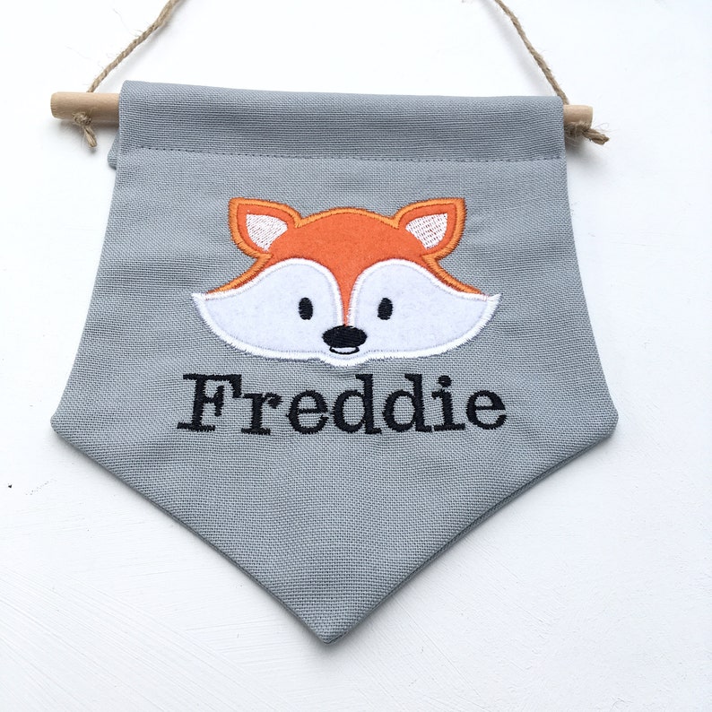May include: A gray fabric banner with a fox design and the name "Freddie" embroidered in black. The fox has orange and white details. The banner is suspended by a wooden dowel and a natural twine.