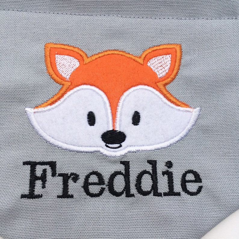 May include: Gray pet bandana with an embroidered orange fox face and the name "Freddie" in black. The fox has white accents and black eyes and nose. The bandana is made of a soft fabric.