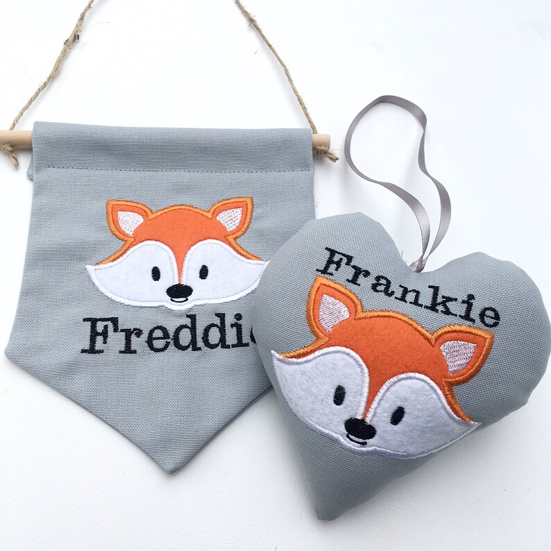 May include: A gray banner and heart-shaped ornament featuring embroidered fox faces. The banner has the name "Freddie" embroidered below the fox. The heart-shaped ornament has the name "Frankie" embroidered above the fox.