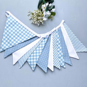 May include: A blue and white fabric bunting featuring a mix of patterns such as gingham, stripes, and polka dots. The triangular flags are connected by a white ribbon. A small bunch of white flowers is in the background.