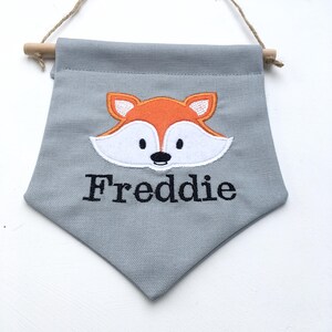 May include: A gray fabric banner with a wooden dowel and twine for hanging. The banner features an orange and white fox face applique and the name "Freddie" embroidered in black.