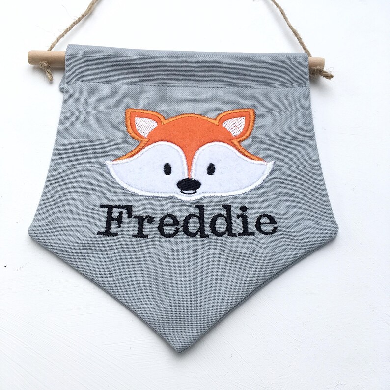 May include: A gray fabric banner with a wooden dowel and twine for hanging. The banner features an orange and white fox face applique and the name "Freddie" embroidered in black.