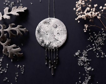 Puede incluir: Un collar con un colgante de luna plateado, con un diseño detallado de la superficie de la luna y acentos de cristal negro que cuelgan de una cadena plateada. El collar está rodeado de pequeños cristales blancos y una rama de flores blancas.