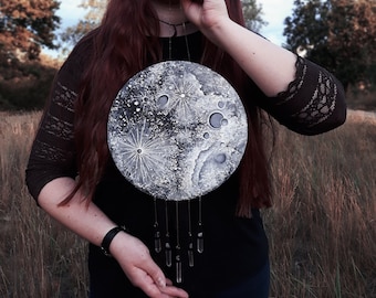 Puede incluir: Una mujer con una blusa negra y un collar con un colgante de luna plateado con detalles de cristal. El colgante de luna tiene un diseño pintado en blanco y gris.