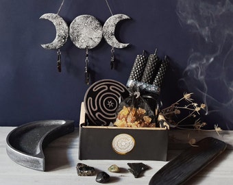 May include: A black wooden box with a black and white moon and star design on the lid. The box is filled with black candles, incense, and crystals. The box is sitting on a wooden table with a black and white moon and star design on the lid.