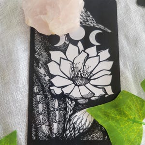 May include: Black and white tarot card with the number XVIII and the words "The Moon" printed on the bottom. The card depicts a large flower with a crescent moon above it.