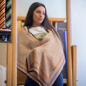 May include: A woman wearing a light brown scarf with a white, orange, and blue patterned border. The scarf is wrapped around her neck and shoulders. She is looking to the right.