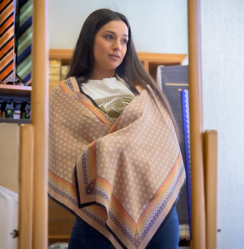 May include: A woman wearing a light brown scarf with a white, orange, and blue patterned border. The scarf is wrapped around her neck and shoulders. She is looking to the right.