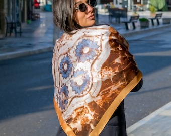 May include: A woman wearing a brown and white silk scarf with a blue floral pattern and gold trim. The scarf is tied around her neck and drapes down her back.