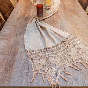 May include: A beige table runner with a crocheted lace trim and fringe. The runner is laid on a wooden table with brown chairs.