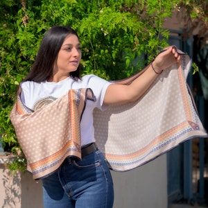 May include: A woman wearing a white t-shirt and blue jeans is holding a beige silk scarf with a polka dot pattern and a colorful border. The scarf is draped over her shoulders and she is holding it up with her right hand.