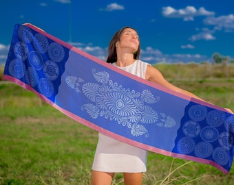 May include: A long, rectangular scarf with a blue background and white decorative patterns. The scarf has a pink border and is being held up against a blue sky and green field.