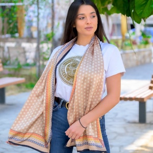 May include: A woman wearing a white t-shirt with a gold logo that says "Versace Jeans" is wearing a beige scarf with white polka dots and a colorful border. The scarf is draped around her neck and shoulders.