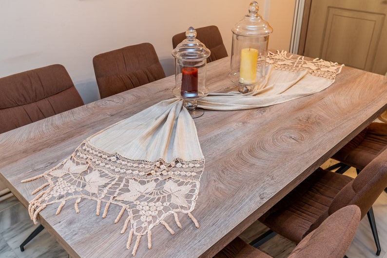 May include: A wooden dining table with brown chairs and a crocheted table runner with a fringe. Two glass jars with candles are on the table runner.
