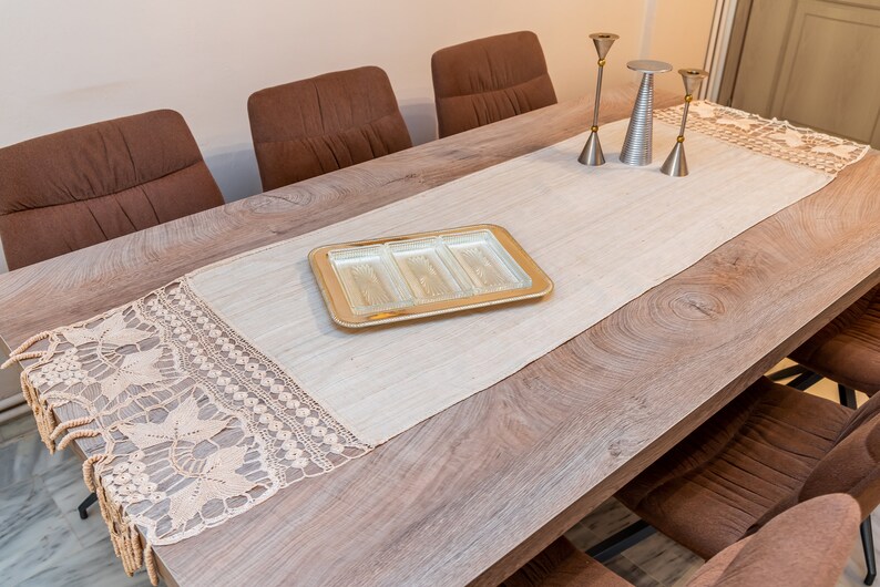 May include: A wooden dining table with a beige table runner and a crocheted lace trim. The table runner has a glass serving tray with four compartments in the center. There are four brown chairs around the table.