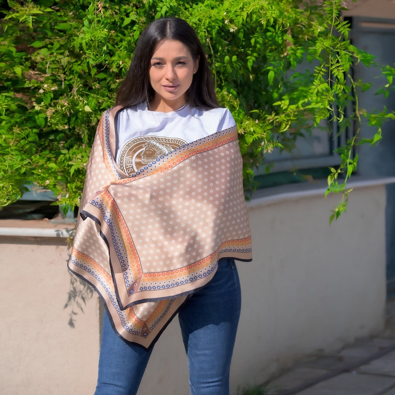 May include: A woman wearing a beige scarf with white polka dots and a black, orange, and blue border. The scarf is tied around her neck and draped over her shoulders. She is wearing a white t-shirt with a gold design and blue jeans.