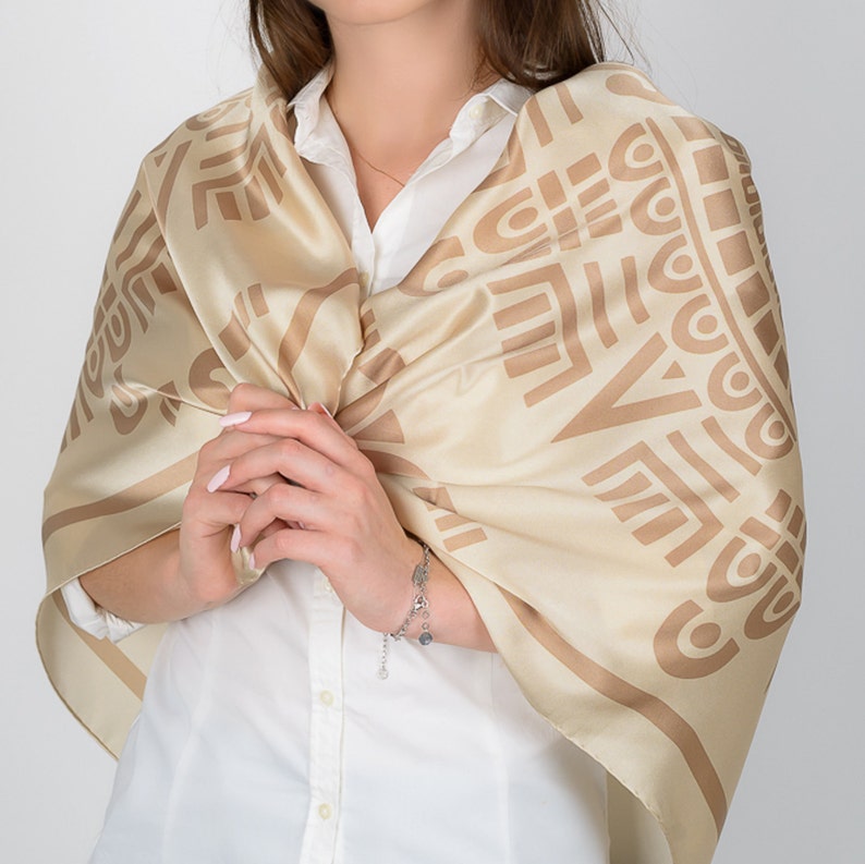 May include: A beige silk scarf with a geometric pattern in a darker shade of beige. The scarf is draped over a white collared shirt. A silver bracelet with charms is visible on the wrist.