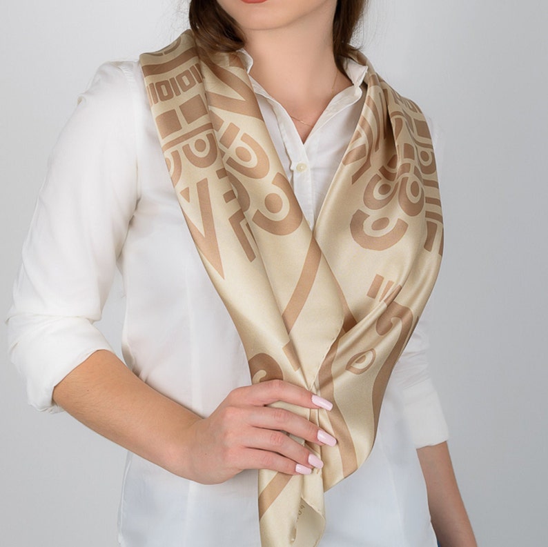 May include: A beige and tan silk scarf with a geometric pattern. The scarf is draped around the neck over a white collared shirt. The scarf's design features repeating abstract shapes and lines.