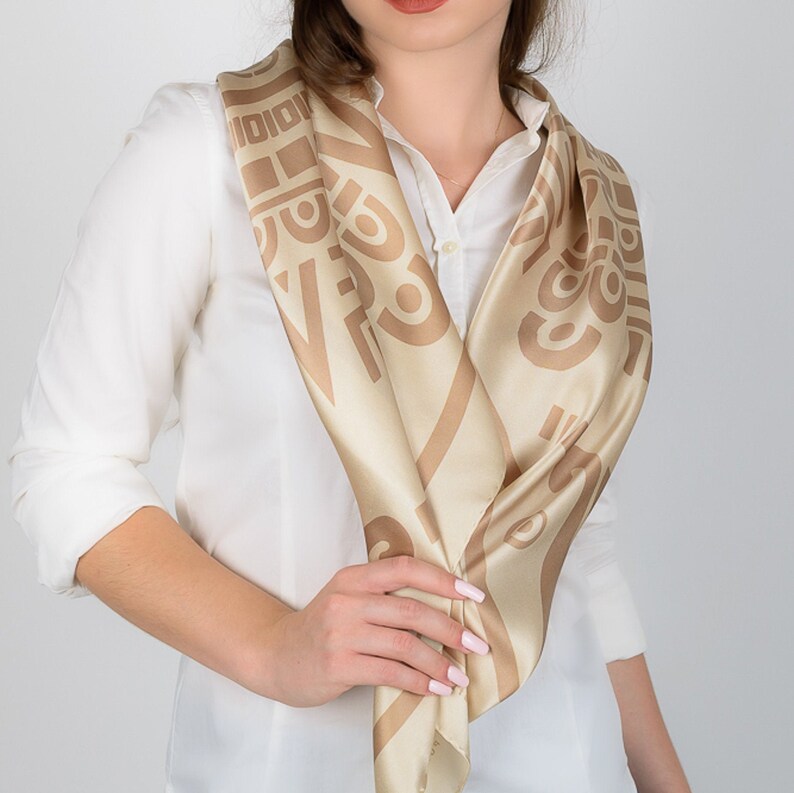 May include: A beige and brown patterned silk scarf draped around the neck. The scarf features a repeating geometric design in a darker shade of brown against a lighter beige background. The scarf is worn over a white collared shirt.
