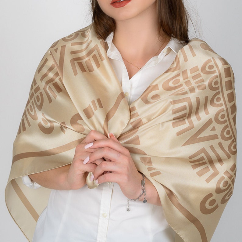 May include: A beige silk scarf with a geometric pattern in a darker shade of beige. The scarf is draped over a white collared shirt. The scarf's design features repeating shapes and lines, creating a textured appearance. The scarf is a square shape.