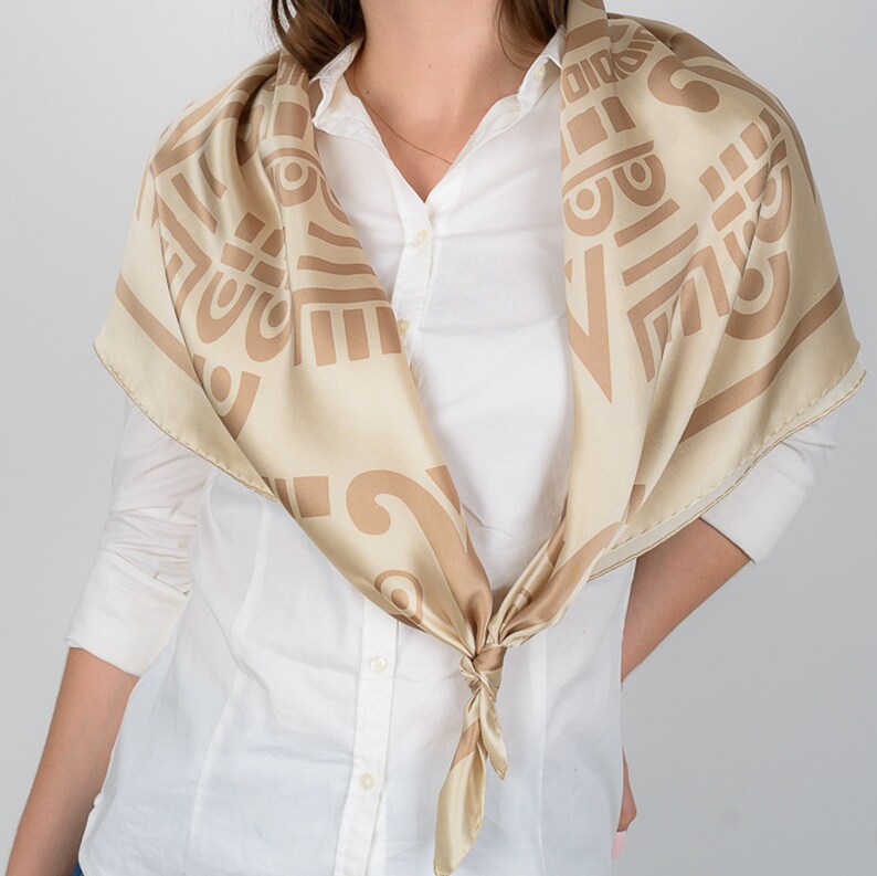 May include: A beige silk scarf with a repeating geometric pattern in a darker beige hue. The scarf is knotted at the front, showcasing the intricate design. The scarf is worn over a white button-down shirt.