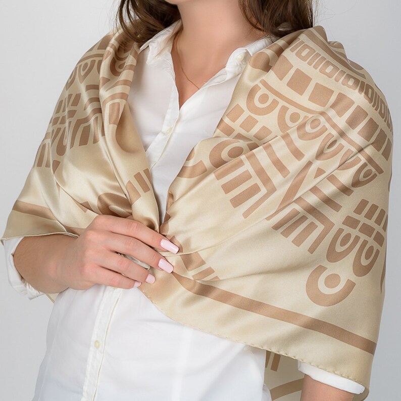 May include: A beige silk scarf with a geometric pattern in a darker shade of beige. The scarf is draped over a white shirt. The pattern consists of squares, rectangles, and curved lines, creating a symmetrical design.